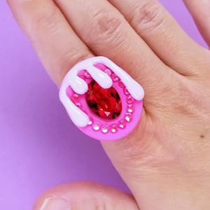 Könnte beinhalten: Ein pinkfarbener Ring mit einem großen, facettierten roten Edelstein in der Mitte, umgeben von kleinen, klaren Kristallen. Der Ring hat ein weißes, tropfendes Eis-Design. Der Ring befindet sich auf einem Finger vor einem lila Hintergrund.