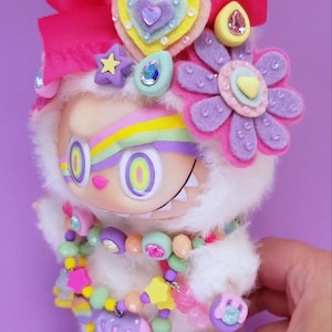 May include: A whimsical, plush doll with a white furry body, adorned with a large pink bow, felt flowers, and a colorful beaded necklace. The doll has large, round eyes and a rainbow-colored stripe across its forehead. The overall design is bright and playful.
