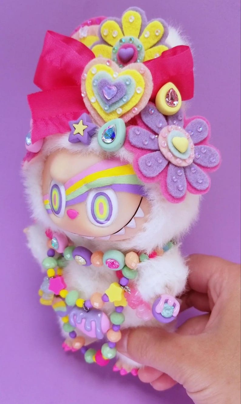 May include: A whimsical, plush doll with a white furry body, adorned with a large pink bow, felt flowers, and a colorful beaded necklace. The doll has large, round eyes and a rainbow-colored stripe across its forehead. The overall design is bright and playful.