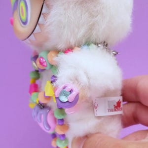 May include: A plush white monster toy with a colorful beaded necklace and a flower crown. The monster has a large, toothy grin and a tag that reads "THEMONS". The toy is held against a purple background.