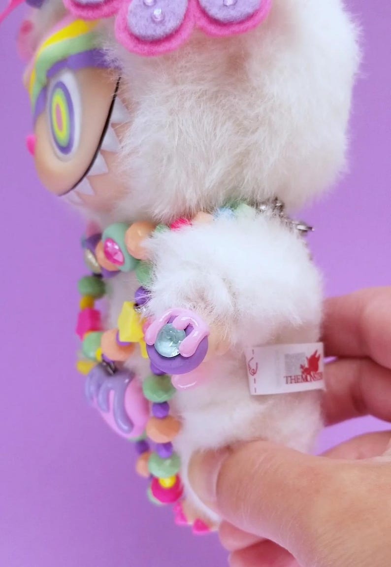 May include: A plush white monster toy with a colorful beaded necklace and a flower crown. The monster has a large, toothy grin and a tag that reads "THEMONS". The toy is held against a purple background.