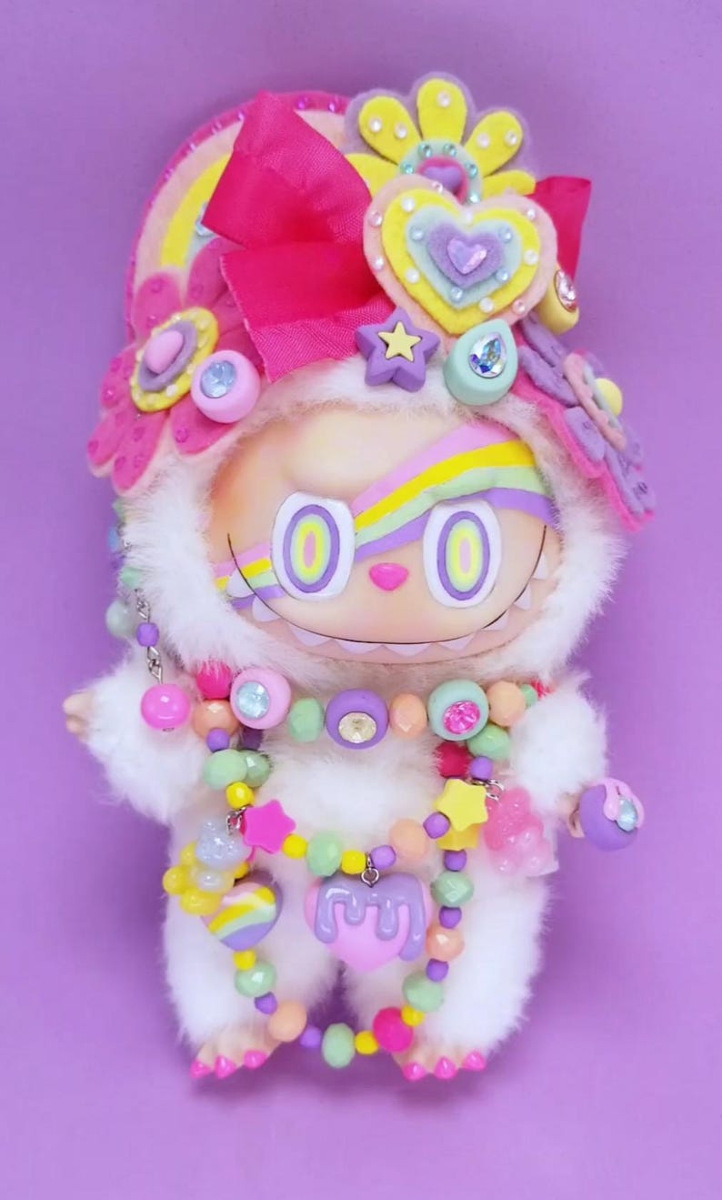 May include: A whimsical plush doll with a white furry body and a smiling face. It is adorned with a rainbow-striped forehead, a pink bow, and a colorful beaded necklace. The doll is decorated with hearts, stars, and flowers, set against a purple background.