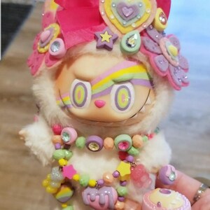 May include: A whimsical doll with a white furry body, adorned with a pink bow, floral and heart-shaped embellishments. It features a colorful rainbow design across the eyes and a beaded necklace. A small pink trinket box is held nearby.