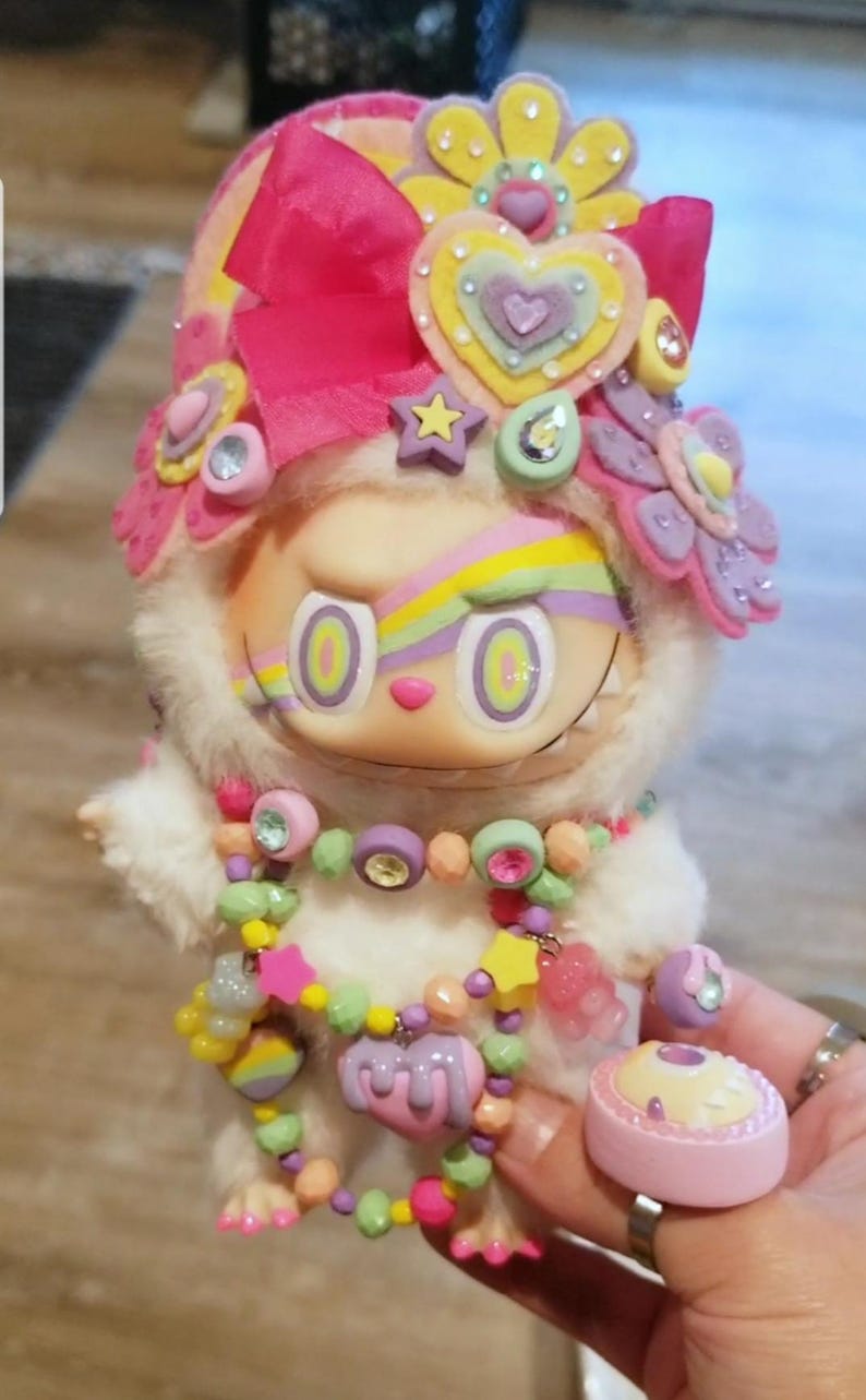 May include: A whimsical doll with a white furry body, adorned with a pink bow, floral and heart-shaped embellishments. It features a colorful rainbow design across the eyes and a beaded necklace. A small pink trinket box is held nearby.