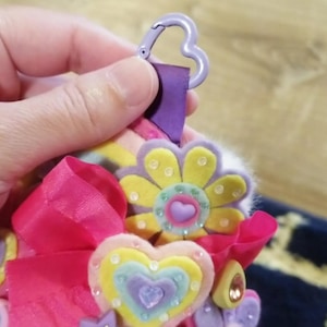 May include: A colorful keychain with felt flower and heart embellishments. The keychain features a purple heart-shaped clasp, a pink ribbon, and various pastel-colored felt shapes. The flowers and hearts are adorned with small, sparkling gems.