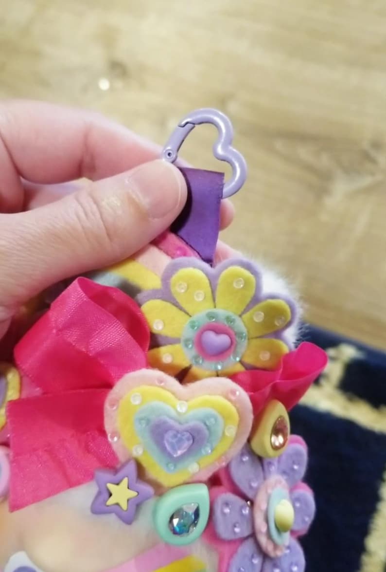 May include: A colorful keychain with felt flower and heart embellishments. The keychain features a purple heart-shaped clasp, a pink ribbon, and various pastel-colored felt shapes. The flowers and hearts are adorned with small, sparkling gems.