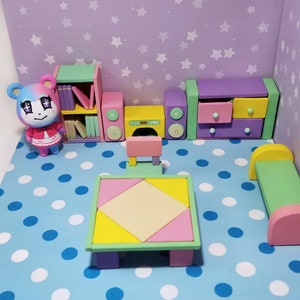 May include: A miniature dollhouse room with a pink and blue polka dot floor. The room features a bookshelf, a stereo system, a table and chairs, a dresser, and a bed. The room is decorated with a light purple wall with white stars.