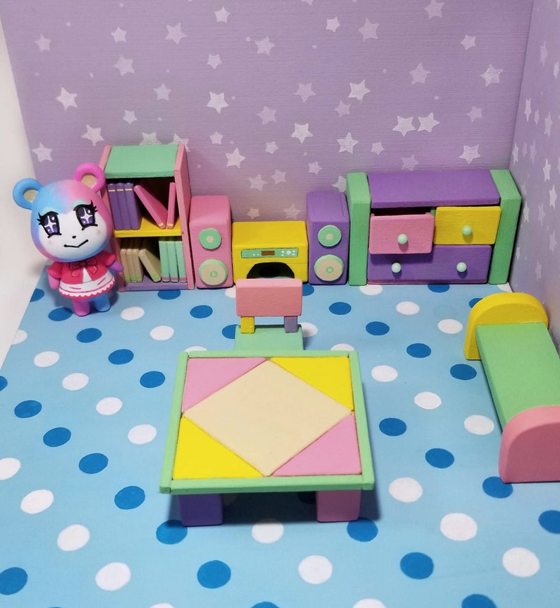 May include: A miniature dollhouse room with a pink and blue polka dot floor. The room features a bookshelf, a stereo system, a table and chairs, a dresser, and a bed. The room is decorated with a light purple wall with white stars.