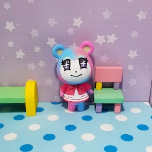 May include: A pink and blue animal character figurine with a white dress and red bow. The figurine is standing on a blue and white polka dot surface.