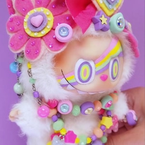 May include: A whimsical, plush doll with a white furry body, adorned with colorful accessories. It features a rainbow headband, pink flower accents, and a beaded necklace. The doll has large, round eyes and a painted smile, creating a playful aesthetic.