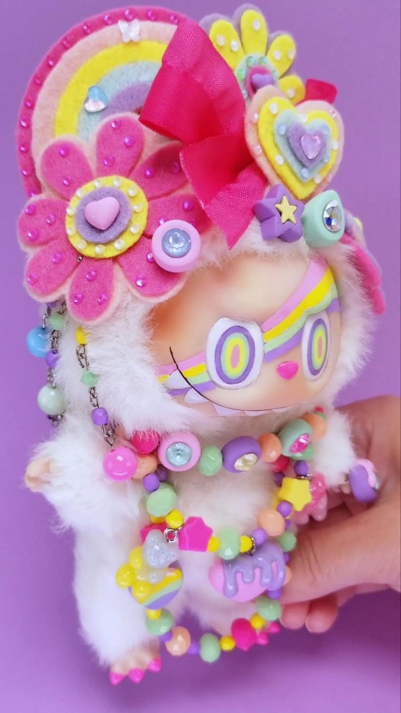 May include: A whimsical, plush doll with a white furry body, adorned with colorful accessories. It features a rainbow headband, pink flower accents, and a beaded necklace. The doll has large, round eyes and a painted smile, creating a playful aesthetic.