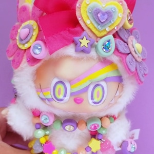 May include: A whimsical doll with a white furry hood and a colorful rainbow design on its face. The doll is adorned with felt flowers, a pink bow, and a beaded necklace with star and heart charms. The overall design is bright and playful.