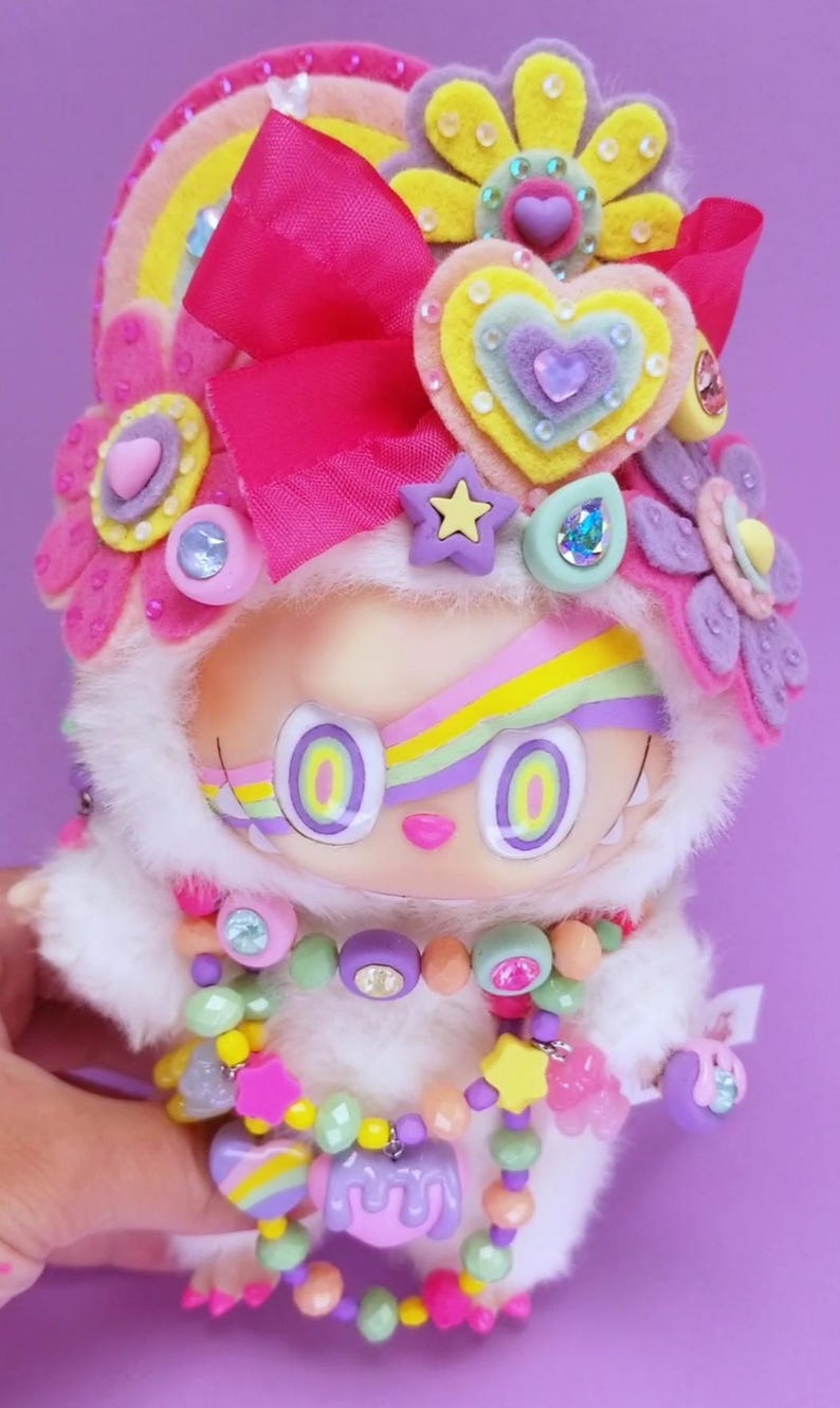 May include: A whimsical doll with a white furry hood and a colorful rainbow design on its face. The doll is adorned with felt flowers, a pink bow, and a beaded necklace with star and heart charms. The overall design is bright and playful.