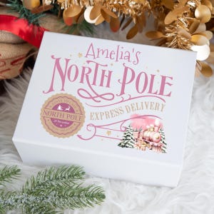 May include: A white gift box with pink text reading "Amelia's North Pole Express Delivery". The box features a train illustration and a North Pole seal. Gold and green decorations surround the box.