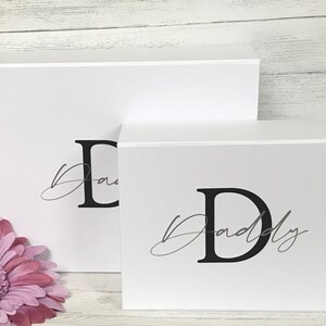 Pu&ograve; includere: Due scatole regalo bianche con coperchi. La scatola pi&ugrave; grande ha la lettera "D" e la parola "Pap&agrave;" scritta in corsivo nero. La scatola pi&ugrave; piccola ha la lettera "D" e la parola "Pap&agrave;" scritta in corsivo nero.