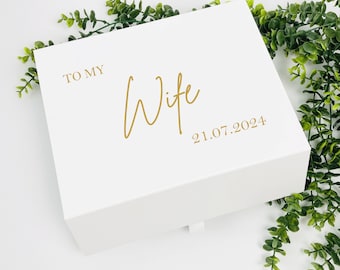 May include: A white gift box with gold lettering that reads "TO MY Wife 21.07.2024". The box is rectangular with a lid and a small ribbon pull. Green foliage is in the background.