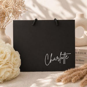 May include: A black gift bag with black rope handles. The name "Charlotte" is written in white cursive script on the front of the bag. The bag is rectangular and sits on a light-colored surface, with decorative flowers and vases in the background.