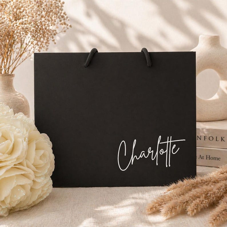 May include: A black gift bag with black rope handles. The name "Charlotte" is written in white cursive script on the front of the bag. The bag is rectangular and sits on a light-colored surface, with decorative flowers and vases in the background.