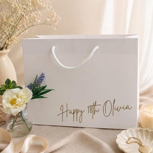 Pode incluir: Uma sacola de presente branca com al&ccedil;a branca e o texto "Happy 18th Olivia" em letras douradas. A sacola &eacute; decorada com lavanda e uma flor branca. Um pequeno vaso com uma flor e uma vela est&atilde;o no fundo.