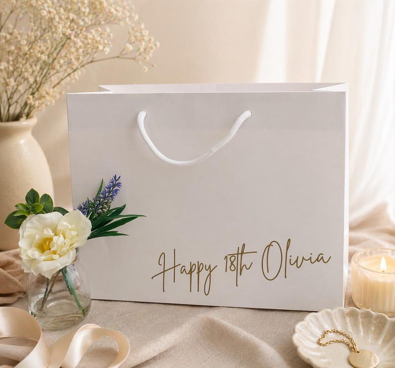 Pode incluir: Uma sacola de presente branca com al&ccedil;a branca e o texto "Happy 18th Olivia" em letras douradas. A sacola &eacute; decorada com lavanda e uma flor branca. Um pequeno vaso com uma flor e uma vela est&atilde;o no fundo.