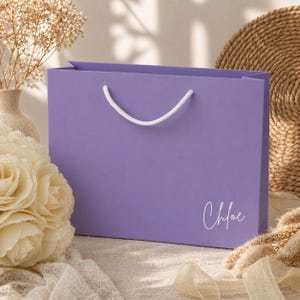 May include: A lavender gift bag with white rope handles. The name "Chloe" is written in cursive on the front. The bag is rectangular and sits on a neutral surface with floral and decorative elements.