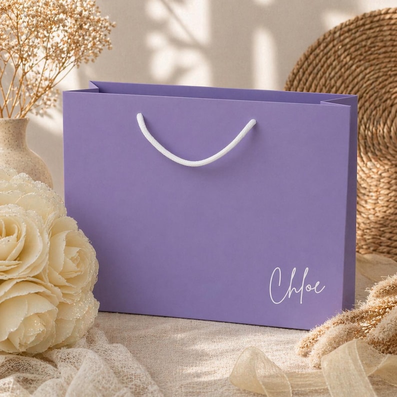 May include: A lavender gift bag with white rope handles. The name "Chloe" is written in cursive on the front. The bag is rectangular and sits on a neutral surface with floral and decorative elements.