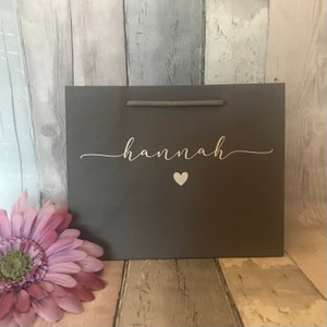 May include: A gray gift bag with a white rope handle and the name "hannah" written in white cursive script with a small white heart below the name.