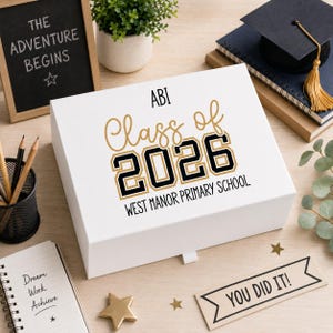 May include: A white gift box with gold and black text reading "ABI Class of 2026" and "West Manor Primary School". The box is on a wooden surface, with a small chalkboard that says "The Adventure Begins".