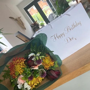 May include: A bouquet of flowers wrapped in green tissue paper with a white gift bag that says "Happy Birthday Dee x". The flowers are a mix of yellow, orange, red, and white.