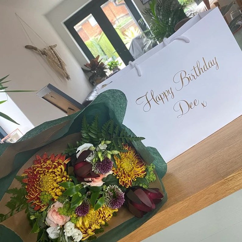 May include: A bouquet of flowers wrapped in green tissue paper with a white gift bag that says "Happy Birthday Dee x". The flowers are a mix of yellow, orange, red, and white.