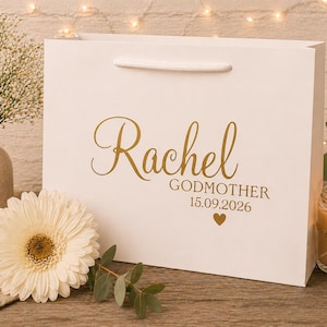 May include: A white gift bag with the name "Rachel" in gold script, along with "Godmother" and the date 15.09.2026. A white flower and a lit candle are next to the bag. The bag has a white handle.