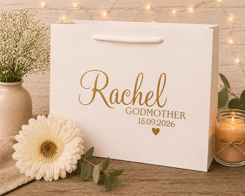 May include: A white gift bag with the name "Rachel" in gold script, along with "Godmother" and the date 15.09.2026. A white flower and a lit candle are next to the bag. The bag has a white handle.
