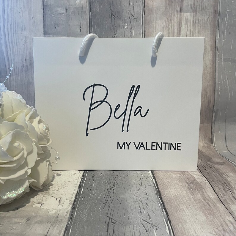 Może przedstawiać: Biała torba prezentowa z czarnymi uchwytami i nadrukiem "Bella My Valentine" na przodzie.