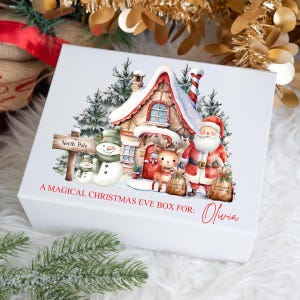 May include: A white box with a Christmas Eve theme. The box features a watercolour illustration of a snowy cottage, Santa Claus, a reindeer, and two snowmen. The text on the box reads "A Magical Christmas Eve Box For: Olivia".
