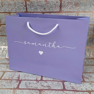 May include: A lavender gift bag with white rope handles. The bag features the name "samantha" in a cursive font, along with a small white heart. The bag is designed for gifting.