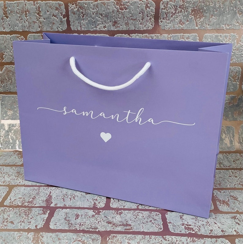 May include: A lavender gift bag with white rope handles. The bag features the name "samantha" in a cursive font, along with a small white heart. The bag is designed for gifting.