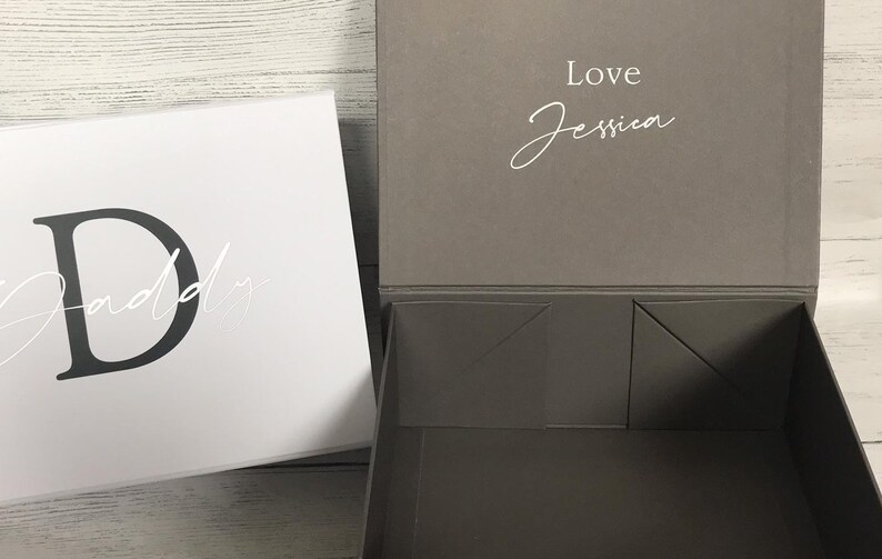Pu&ograve; includere: Una scatola regalo bianca con una lettera nera "D" e la parola "Daddy" scritta in corsivo. Il coperchio della scatola &egrave; grigio e ha le parole "Love Jessica" scritte in corsivo bianco.