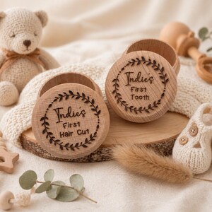 May include: Wooden keepsake boxes with engraved text and a leaf design. One box reads "Indie's First Hair Cut" and the other reads "Indie's First Tooth." A crocheted teddy bear, rattle, and baby booties are also in the image.