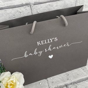 K&ouml;nnte beinhalten: Eine graue Papiert&uuml;te mit wei&szlig;en Henkeln und dem Aufdruck "Kelly's baby shower" auf der Vorderseite.
