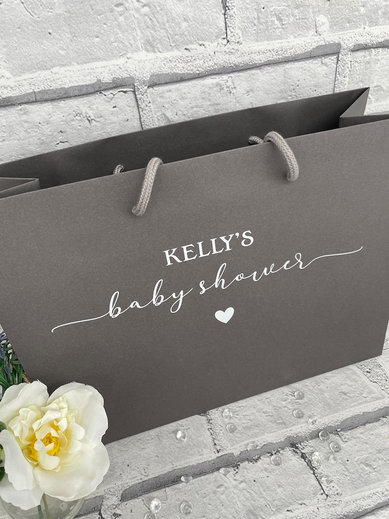 K&ouml;nnte beinhalten: Eine graue Papiert&uuml;te mit wei&szlig;en Henkeln und dem Aufdruck "Kelly's baby shower" auf der Vorderseite.