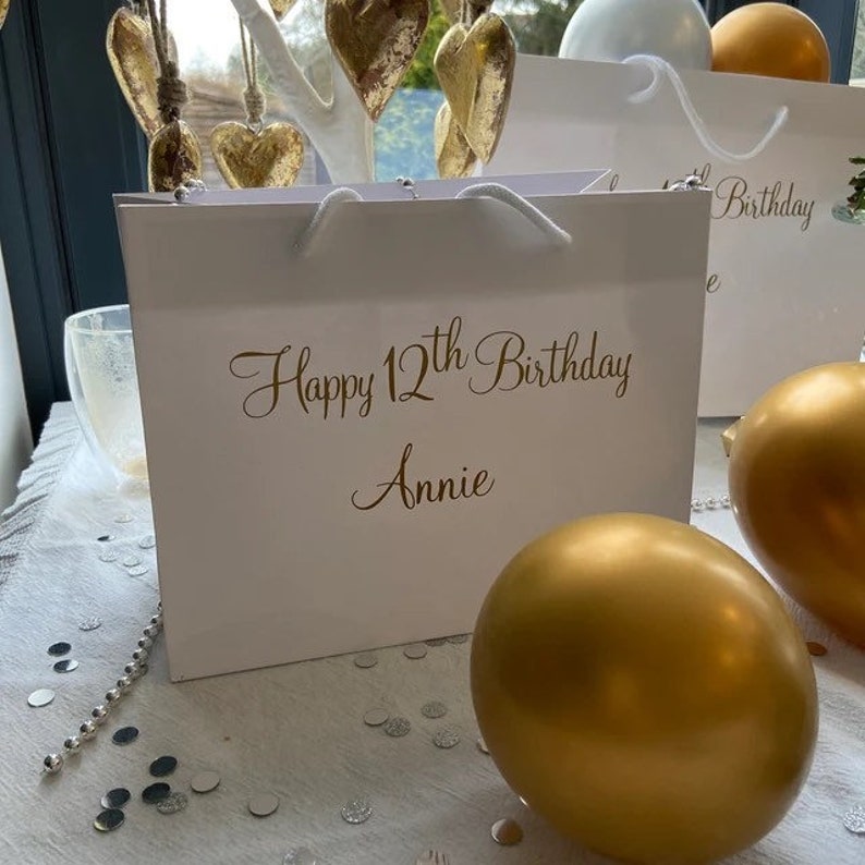 May include: White gift bag with gold lettering that says "Happy 12th Birthday Annie". The bag is sitting on a white table with gold confetti and gold balloons.