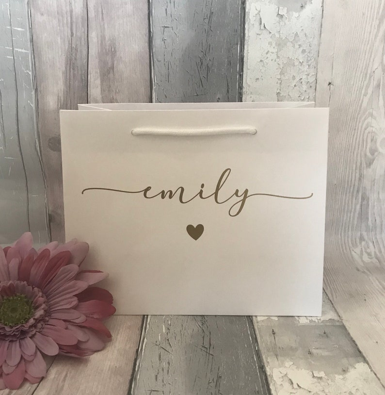 May include: A white gift bag with the name "emily" in gold script and a small gold heart. The bag has a white rope handle and is set against a weathered wood background. A pink flower is in the lower left corner.