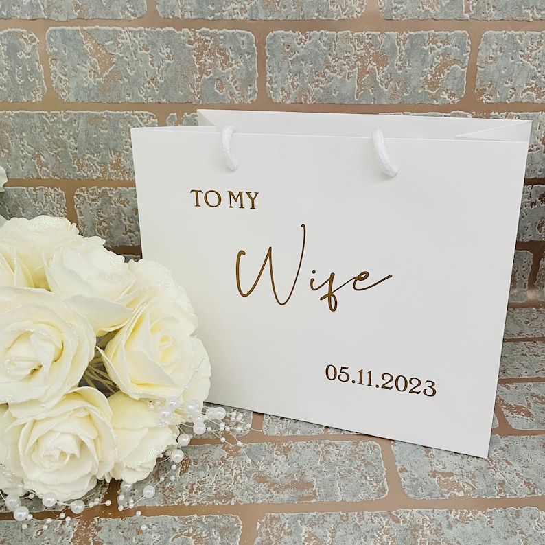 May include: White gift bag with gold lettering that says "To My Wife" and the date "05.11.2023".