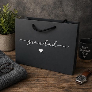 May include: A black gift bag with the word "grandad" in white script and a white heart. A black mug with "Best Grandad Ever" is visible. A watch and a gray sweater are also on the wooden surface.