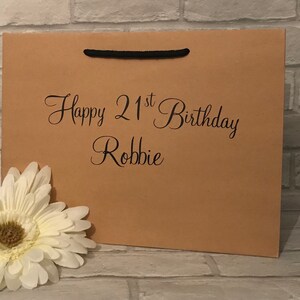 May include: A brown paper gift bag with black handles. The bag has the text "Happy 21st Birthday Robbie" printed on the front.