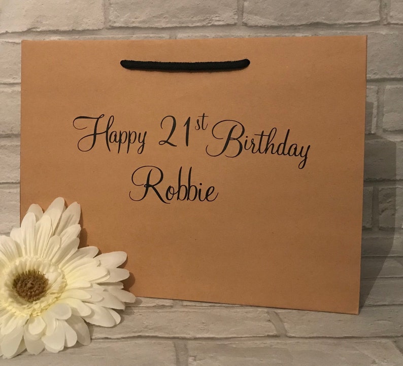 May include: A brown paper gift bag with black handles. The bag has the text "Happy 21st Birthday Robbie" printed on the front.