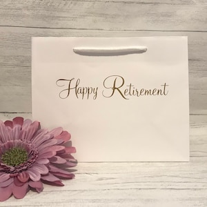 May include: White gift bag with a white rope handle and the text "Happy Retirement" printed in gold.