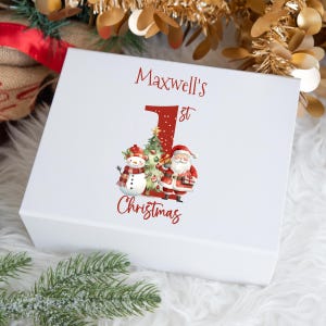 May include: White gift box with a red and green Christmas design. The box says "Maxwell's 1st Christmas" with a snowman and Santa Claus.