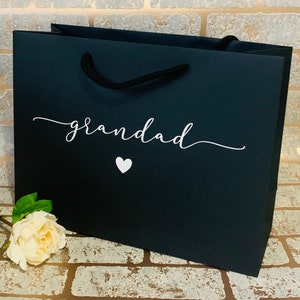 May include: A black gift bag with the word "grandad" in white script and a white heart. The bag has black rope handles and a white flower is placed near the bottom left corner. The bag is sitting on a brick surface.