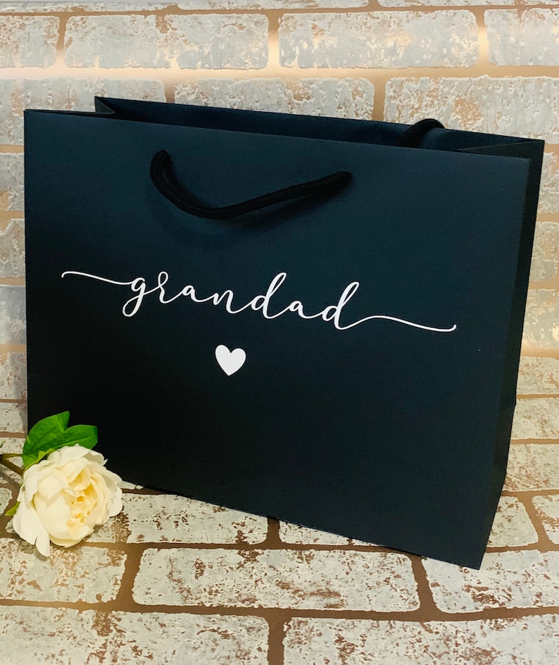 May include: A black gift bag with the word "grandad" in white script and a white heart. The bag has black rope handles and a white flower is placed near the bottom left corner. The bag is sitting on a brick surface.